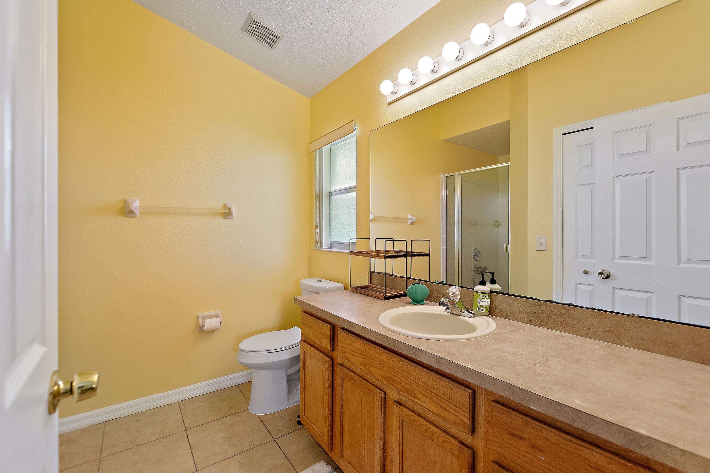 Une salle de bain avec un mur jaune, des toilettes blanches, un meuble-lavabo en bois et un grand miroir avec des lumières au-dessus.