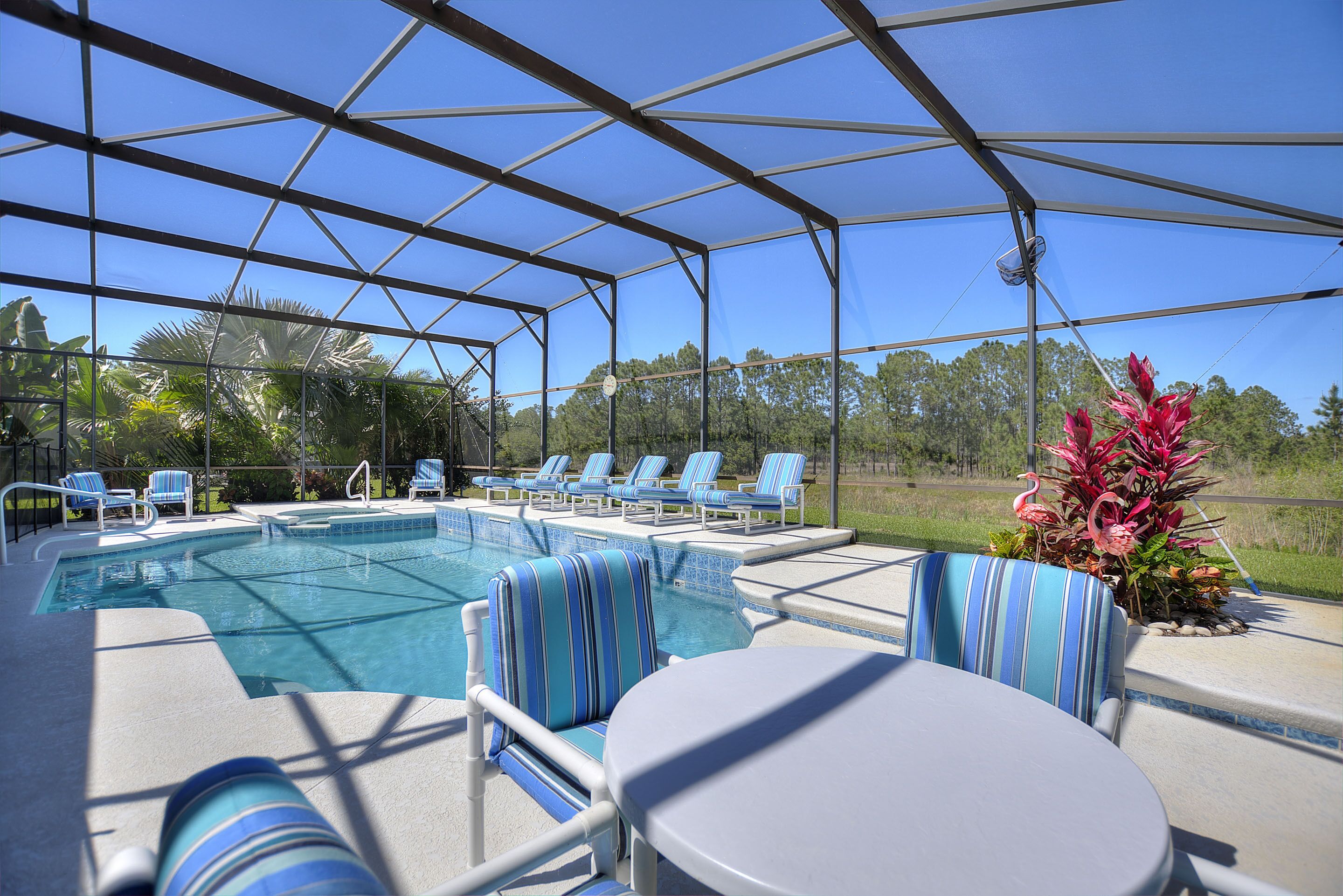 Un área de piscina al aire libre techada con sillones de descanso y una mesa, rodeada por una estructura metálica y paneles de vidrio.