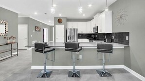 A modern kitchen with three bar stools, a refrigerator, and a microwave. - Jurassic Water View @storey Lake by Shine Villas! Sleeps up to 24! #021 (Kissimmee)