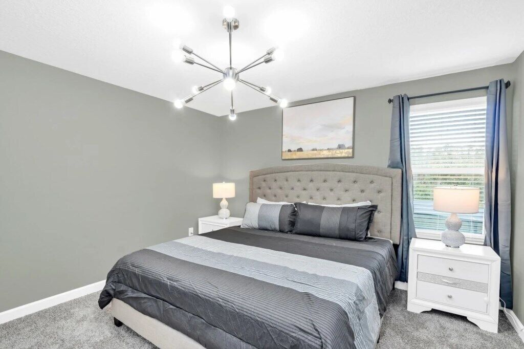 A bedroom with a tufted headboard, a large bed with striped bedding, a bedside table with a lamp, and a window with curtains.