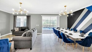 A modern dining area with a long table set for six, blue chairs, and a contemporary chandelier. - Jurassic Water View @storey Lake by Shine Villas! Sleeps up to 24! #021 (Kissimmee)
