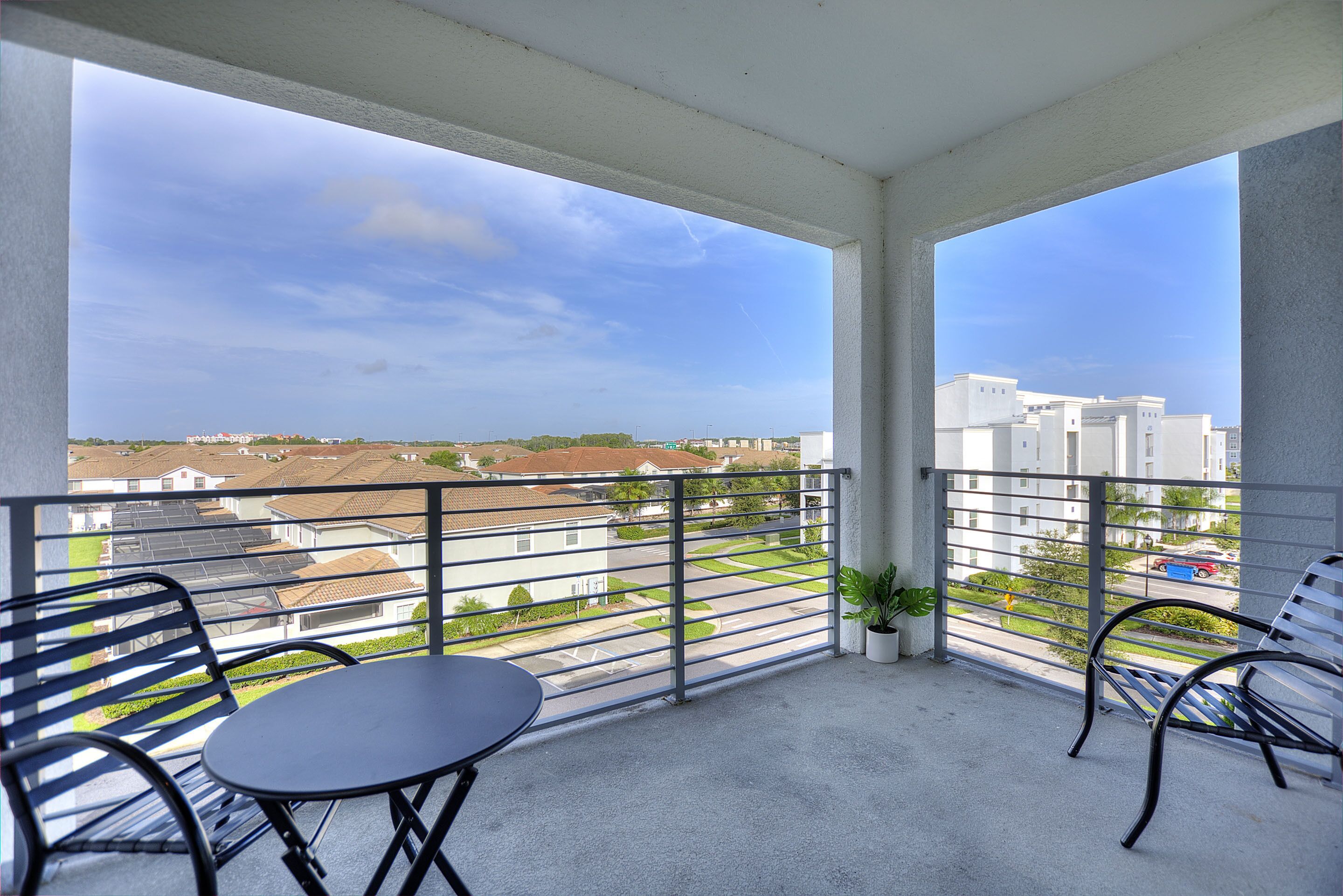 A balcony with a table and chairs, a potted plant, and a view of a residential area with houses and a clear sky.