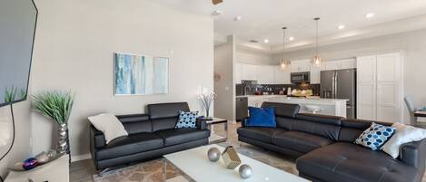 A modern living room with two black sofas, a white coffee table, and a kitchen in the background.