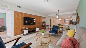 A modern living room with a flat-screen TV, wooden paneling, a round coffee table with a plant, a sofa with colorful cushions, and a dining area with a kitchen in the background.