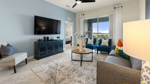 A modern living room with a flat-screen TV, a ceiling fan, a blue sofa, a coffee table, and a view through a large window.