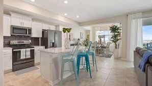Une cuisine moderne avec des meubles blancs, un îlot central, des tabourets de bar de couleur turquoise et une vue sur la salle à manger.