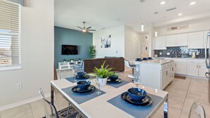 Une cuisine moderne avec des meubles blancs, un coin repas avec une table dressée pour quatre personnes, et un salon avec un canapé marron et un téléviseur fixé au mur.