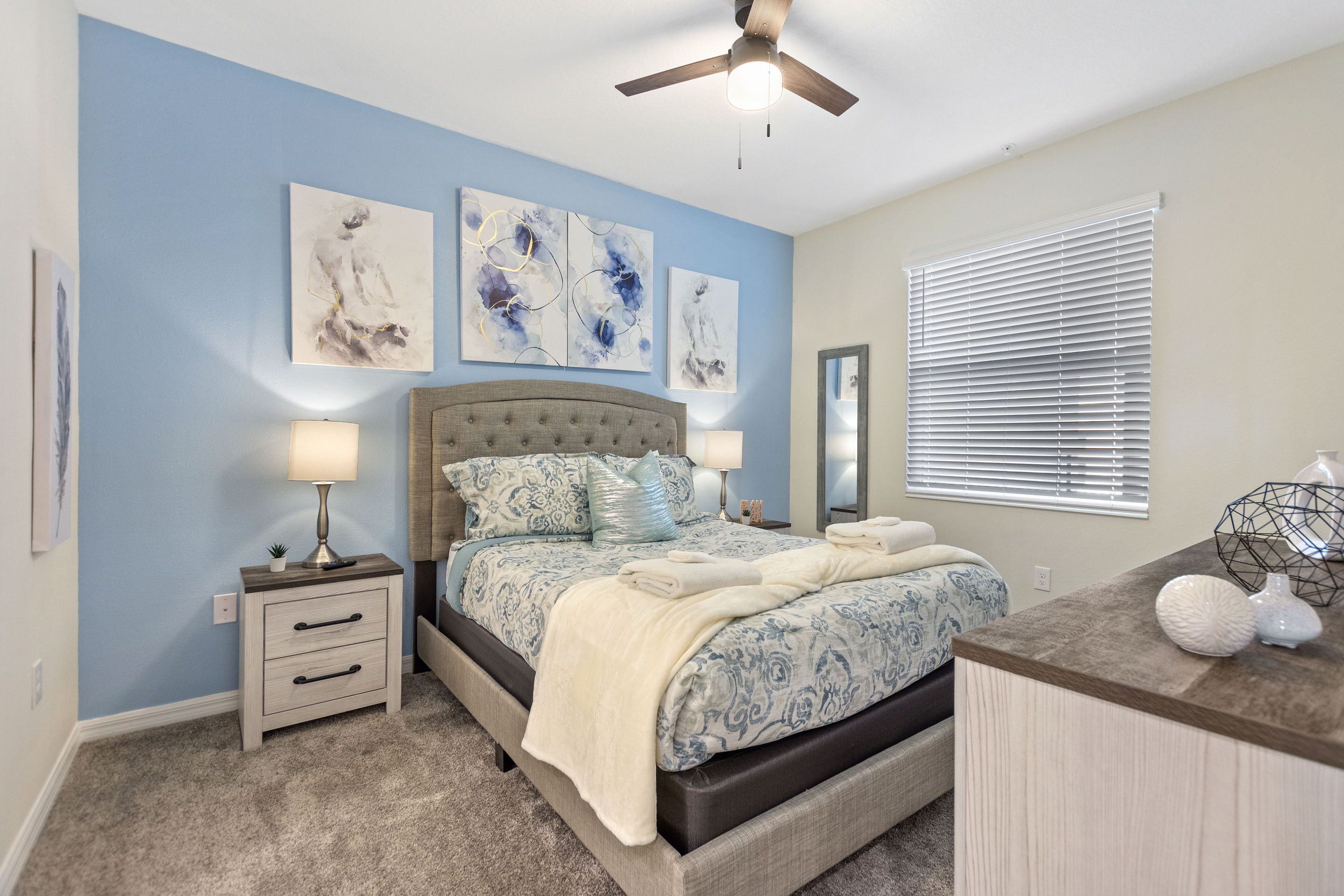 A bedroom with a bed, nightstands, a ceiling fan, artwork on the walls, and a window with blinds.