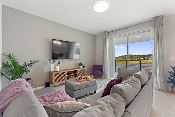 Una sala moderna con un sofá gris, un mueble de entretenimiento de madera, un televisor de pantalla plana y un amplio ventanal corredera que da a un balcón con vistas al paisaje.