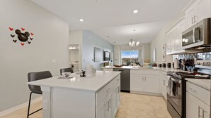 A modern kitchen with white cabinetry, a central island, and a dining area with a chair. - Edgewater Escape at Storey Lake by Shine Villas #904 (Kissimmee)
