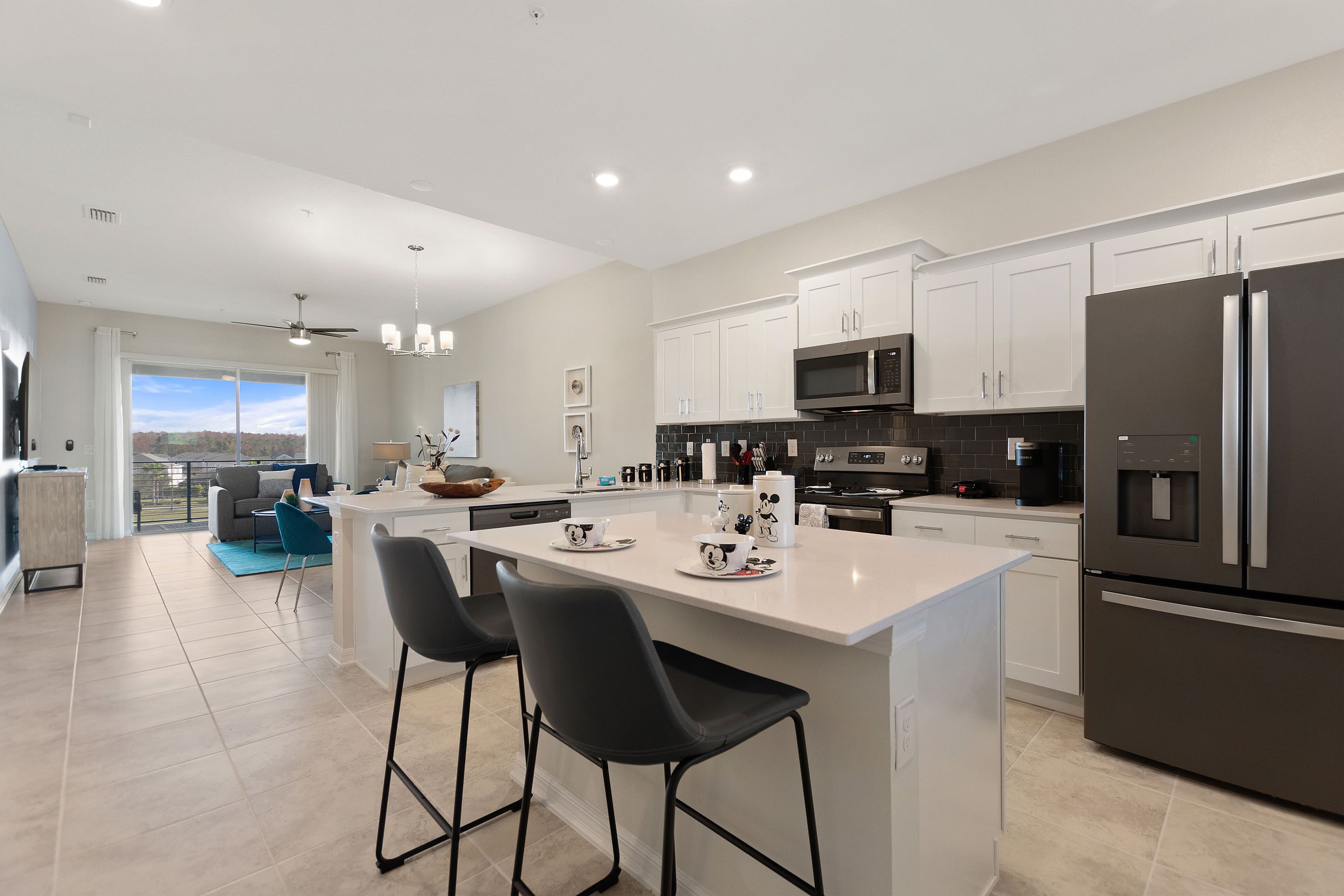 A modern kitchen with a large island, black bar stools, white cabinets, and a stainless steel refrigerator.