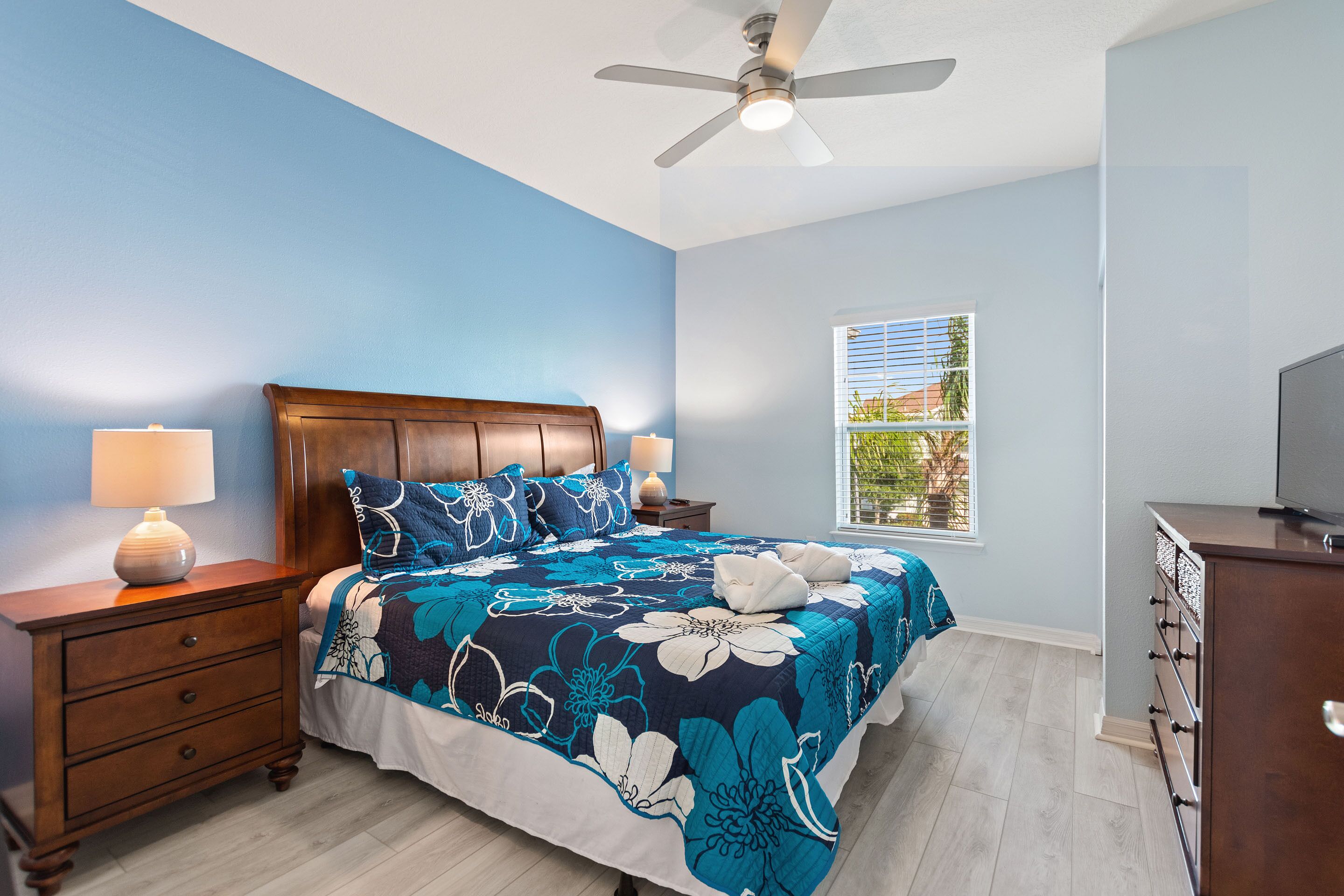 Une chambre à coucher comprenant un lit, des tables de chevet, une commode, un ventilateur de plafond et une fenêtre avec des stores.
