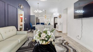 A modern living room with a beige sofa, a black coffee table, and a floral centerpiece. There is a large flat-screen TV mounted on the wall and a chandelier hanging from the ceiling. - Breathe Easy at Solterra Resort by Shine Villas 706 (Davenport)