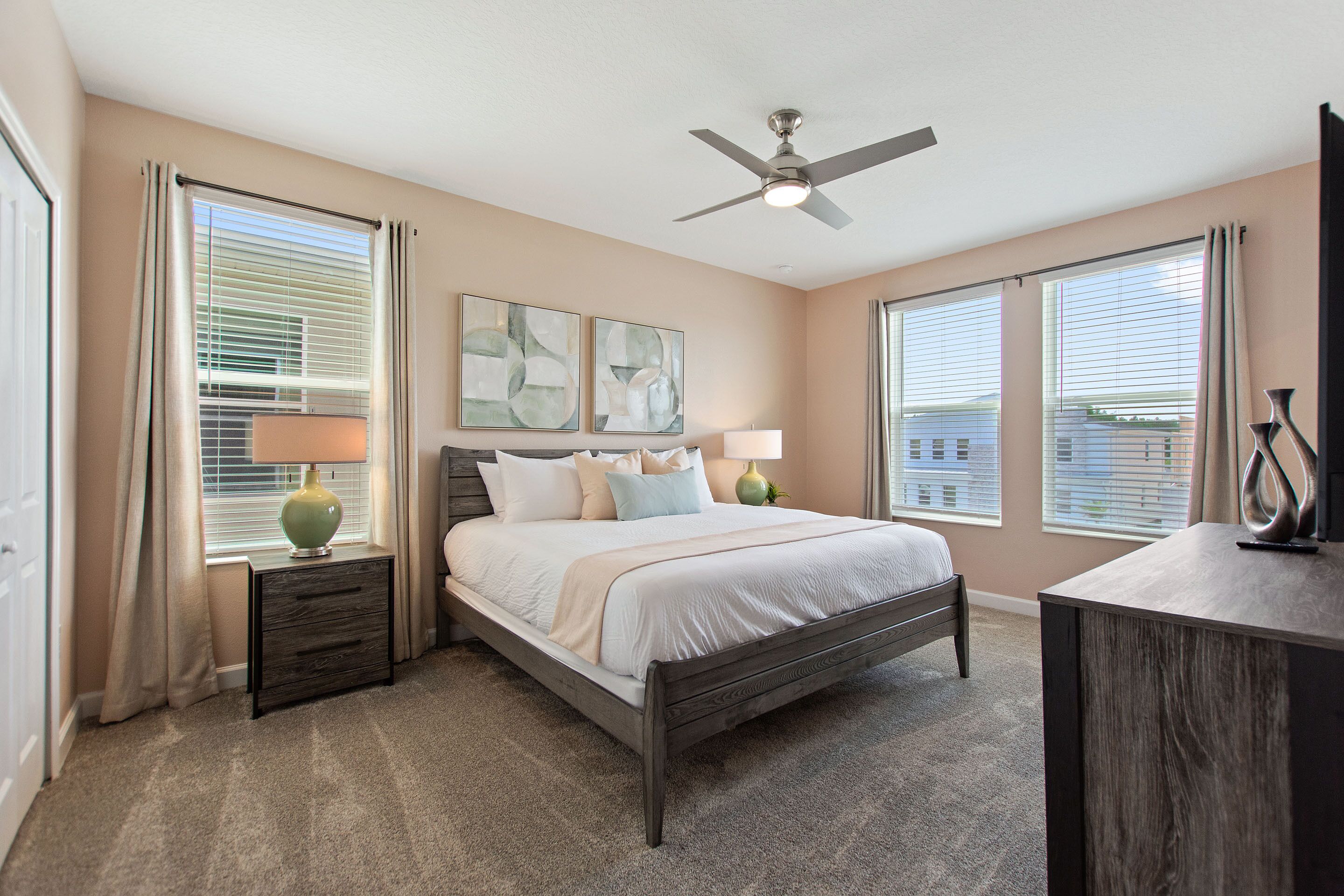 A bedroom with a bed, bedside tables, a ceiling fan, and large windows with blinds.