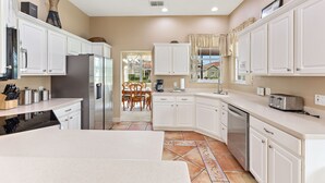 A modern kitchen with white cabinetry, a stainless steel refrigerator, a dishwasher, and a dining area with a table and chairs. - 6 Bedroom Premium Home With Pool, Spa and Games Room #800 (Davenport)