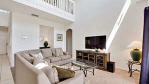 A spacious living room with a high ceiling, a balcony, a flat-screen TV, and a glass coffee table.