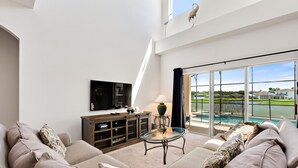 A spacious living room with a large sofa, a glass coffee table, and a TV mounted on the wall. There is a sliding glass door leading to a pool area.