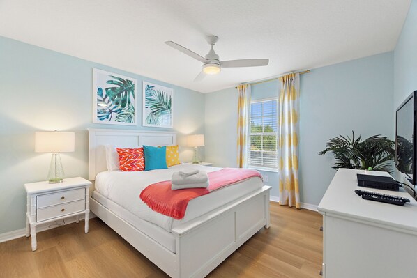 A bedroom with a bed, nightstands, a desk, a ceiling fan, and artwork on the wall.