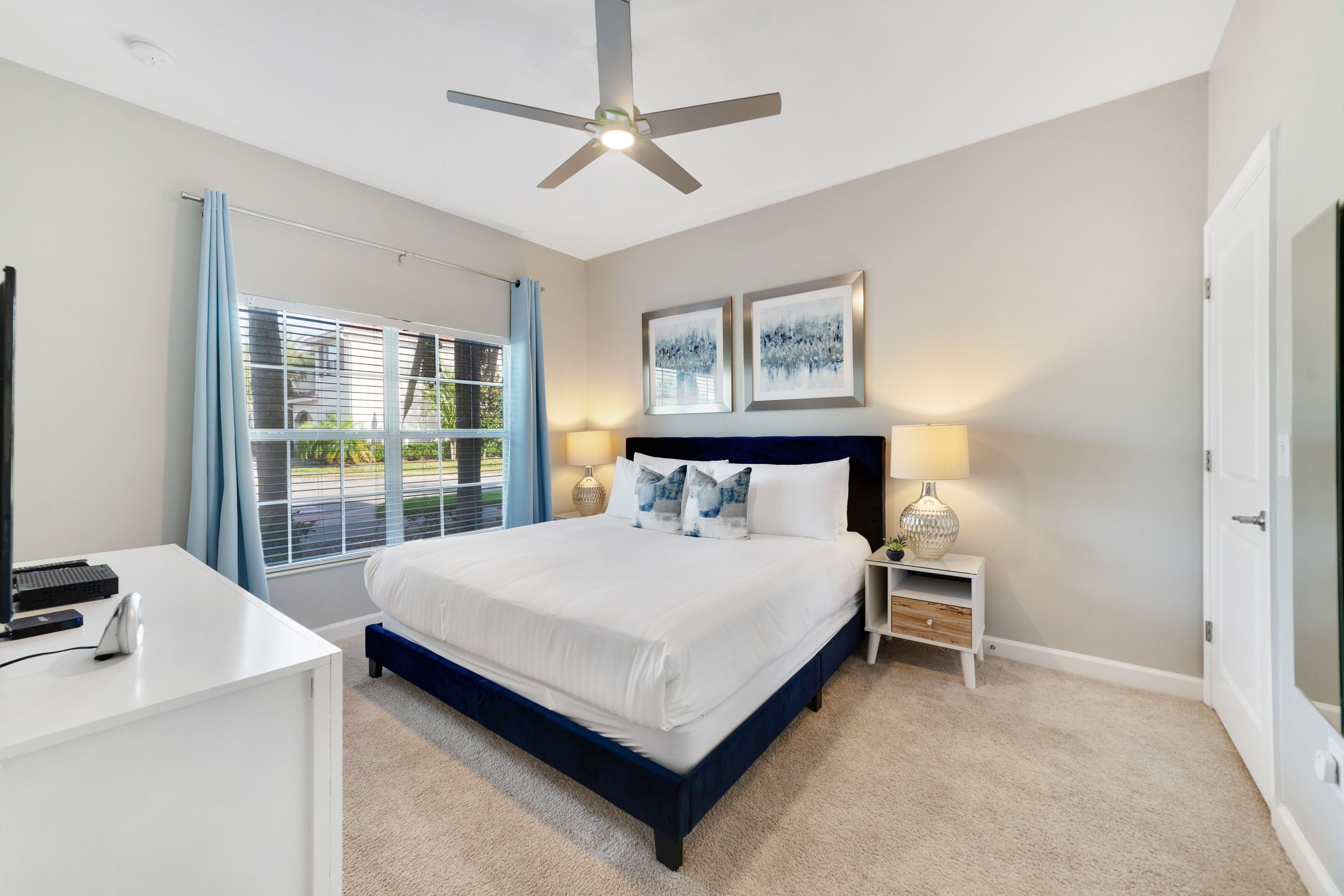A bedroom with a bed, a desk, a ceiling fan, two bedside lamps, and a window with curtains.