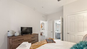 A bedroom with a bed, a dresser, a television, and a bathroom visible through an open door. - Contemporary @vista Cay By Shine Villas #403 (Orlando)