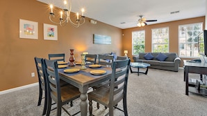 Un coin repas avec une table en bois dressée pour quatre personnes, entourée de chaises. Un lustre est suspendu au-dessus de la table, et un ventilateur de plafond est visible au fond.