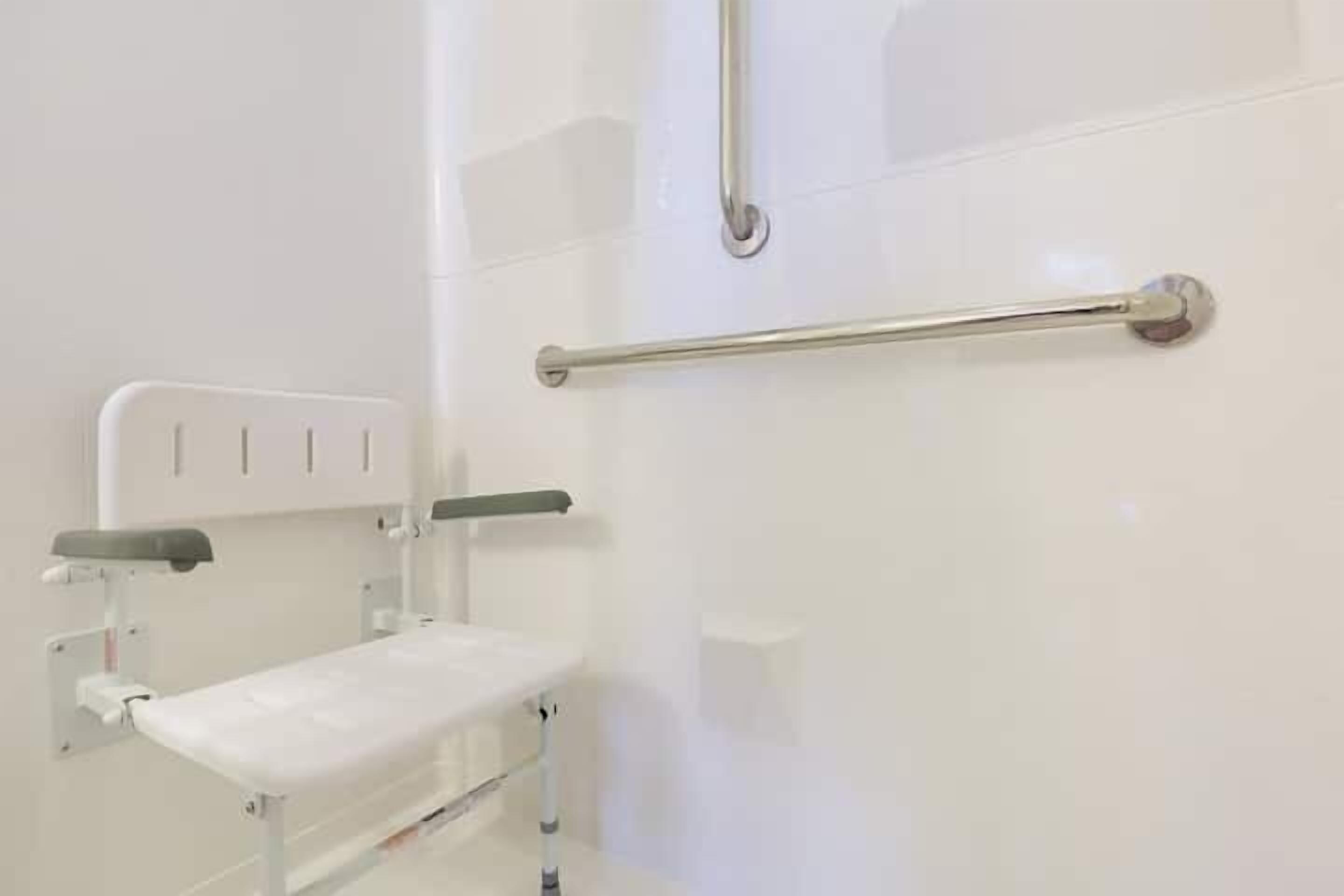 A white bathroom with a white bench, a handrail, and a wall-mounted toilet.