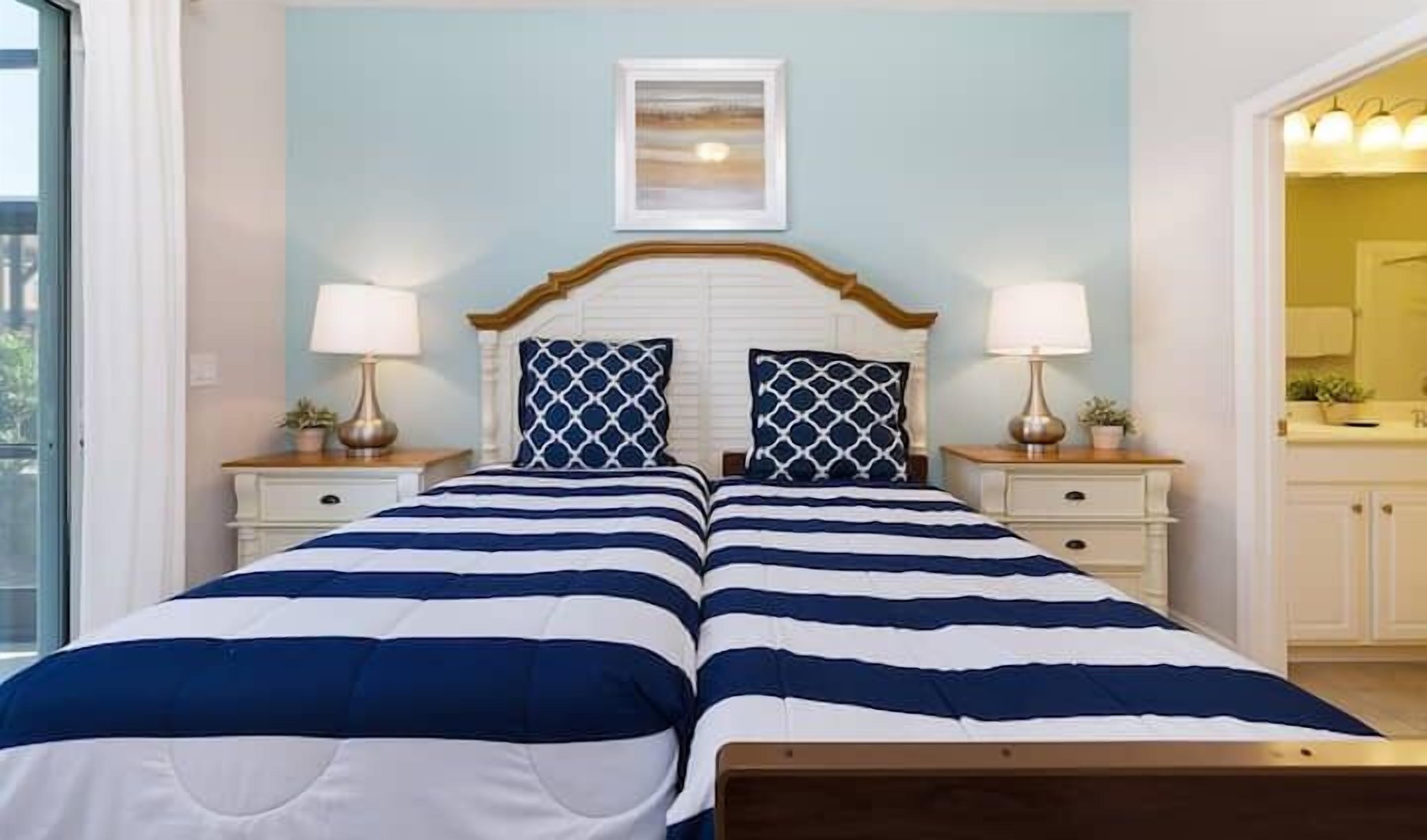 A double bed with a striped bedspread, two bedside tables with lamps, a framed picture above the bed, and a view of a bathroom to the right.