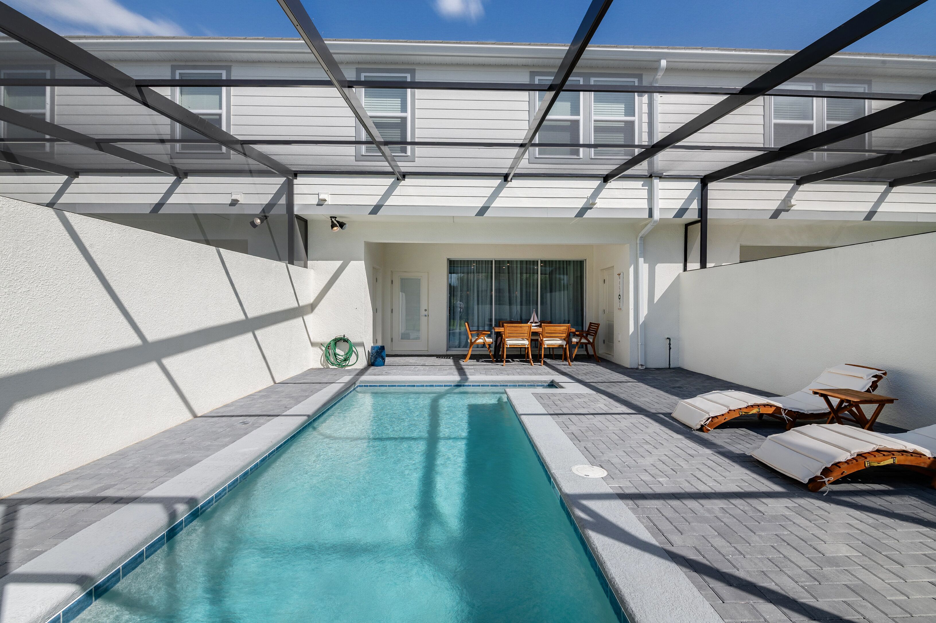 A swimming pool with lounge chairs and a table, surrounded by a white exterior wall and a covered patio area.