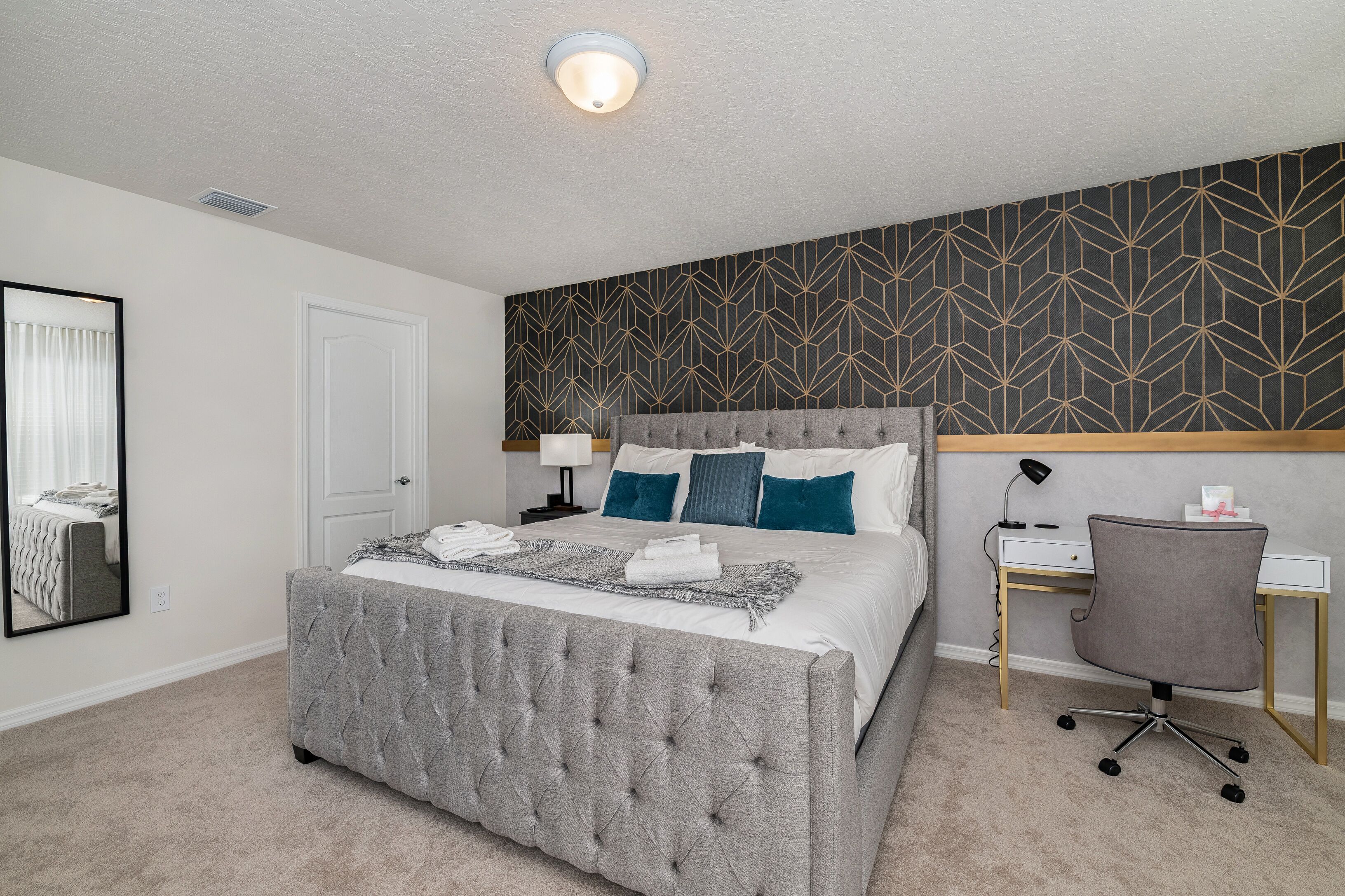 A bedroom with a tufted bed, a desk with a chair, a mirror, and a patterned wall.
