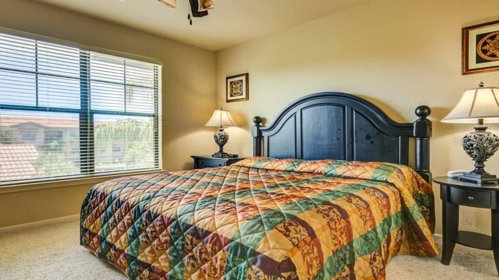 A bedroom with a large bed, a window with blinds, a bedside table with a lamp, and a framed picture on the wall.