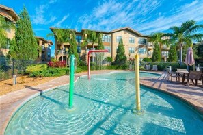 A swimming pool with a water feature, surrounded by a deck with lounge chairs and a view of multi-story buildings. - Sunnyside Up at Bella Piazza by Shine Villas (Davenport)