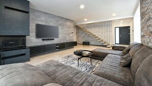 A modern living room with a large flat-screen TV mounted on the wall, a built-in entertainment unit, a grey sectional sofa, a coffee table, and a staircase leading to an upper level. - Stunning home in Vinogradi Ludbreski with sauna (Vinogradi Ludbreski)