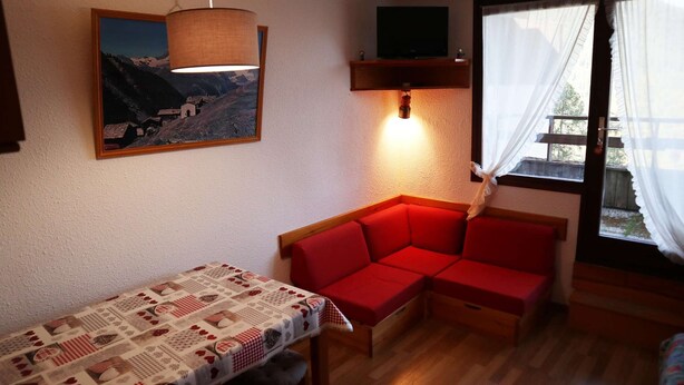 A cozy room with a red sofa, a table with a patterned tablecloth, a painting of a mountain scene, a hanging lamp, and a window with curtains. - STUDIO N° 0309 LE CAIRN (LES ORRES)