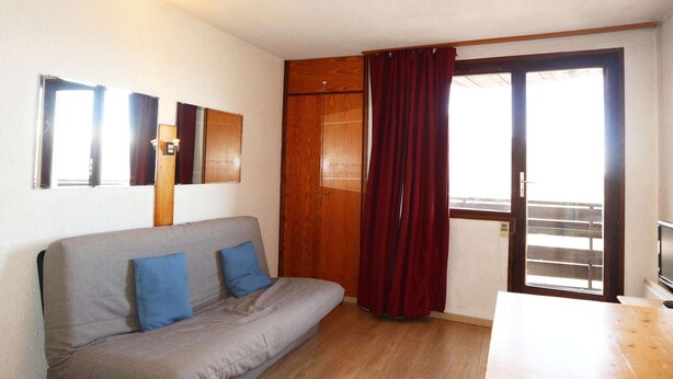 A room with a grey sofa, two blue cushions, a wooden wardrobe, a red curtain, a wooden door, and a television on a wooden table. - COEUR STATION LES ORRES - Res. CAIRN - Studio 4 pers (LES ORRES 1650)