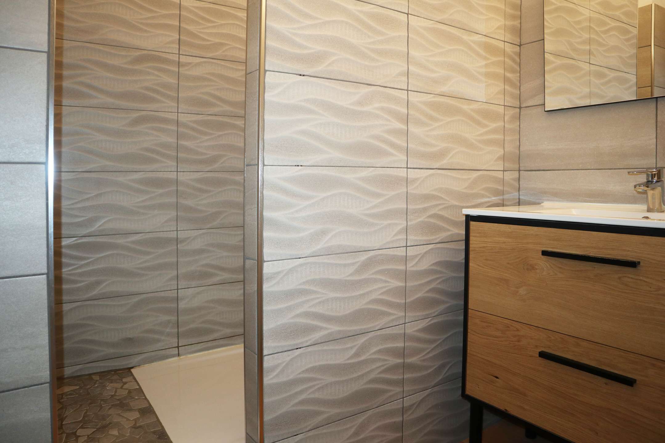 Un bagno con una parete rivestita di piastrelle decorate, un mobile bagno in legno con due cassetti e uno specchio sopra al mobile.