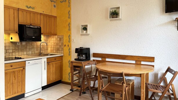 Een compacte keuken met houten kastjes, een magnetron, een koffiezetapparaat en een eettafel met stoelen.