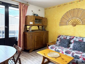 A room with a wooden cabinet, a sofa with a patterned cover, a fan, and a dining table with chairs.