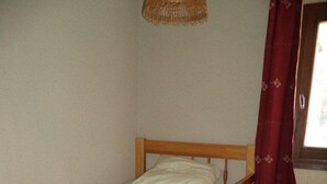 Una cama litera de madera con colchón y almohadas, un taburete de madera con una lámpara, y una ventana con cortina roja.