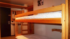 Una cama litera con estructura de madera y ropa de cama blanca. Se observa una puerta y un pequeño estante con un cuadro en la pared.