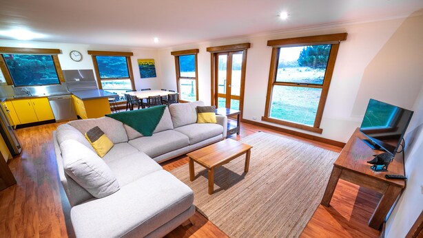 A modern living room with a sofa, coffee table, and TV. There is a kitchen area with a stove and sink, and large windows offering views of the outdoors. - Farm House Accommodation Northern end of the Bay of Fires 5 mins from Mt Willian (Musselroe Bay)