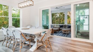 A dining area with a white table and wicker chairs, large windows, and a view of the outdoors. - Newly built 6 bedroom home located at the end of a cul-de-sac for privacy. Large screened in porch (Bald Head Island)
