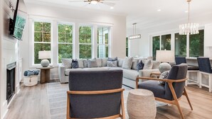 A modern living room with a fireplace, large windows, and a ceiling fan. - Newly built 6 bedroom home located at the end of a cul-de-sac for privacy. Large screened in porch (Bald Head Island)