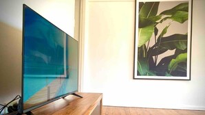 A flat-screen TV on a wooden stand in a room with wooden flooring and a framed plant on the wall. - Homely Vibes 3BR Garden (Padstow)