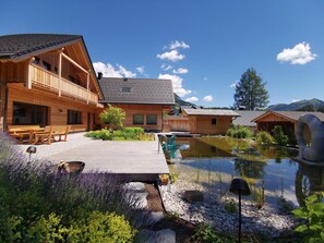 A wooden lodge with a wooden deck, a small pond, and surrounding greenery. - Premium Chalet in Tauplitz With Sauna and Pool (Bad Mitterndorf)