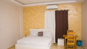 A bedroom with a bed, a desk, a chair, an air conditioner, and a trash bin. - Luxury Palace (Lomé)