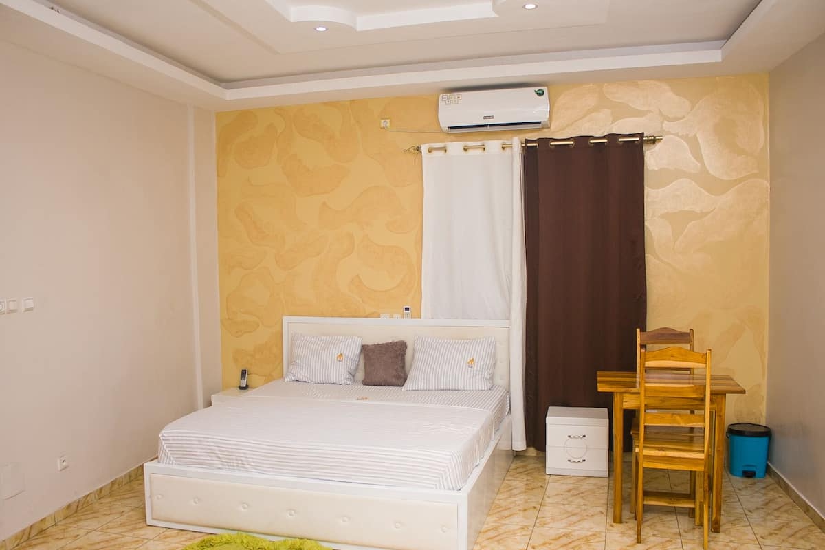 A bedroom with a bed, a desk, a chair, an air conditioner, and a trash bin.