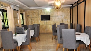 A restaurant with marble walls, a flat-screen TV, and grey upholstered chairs around tables with white tablecloths. - Luxury Palace (Lomé)