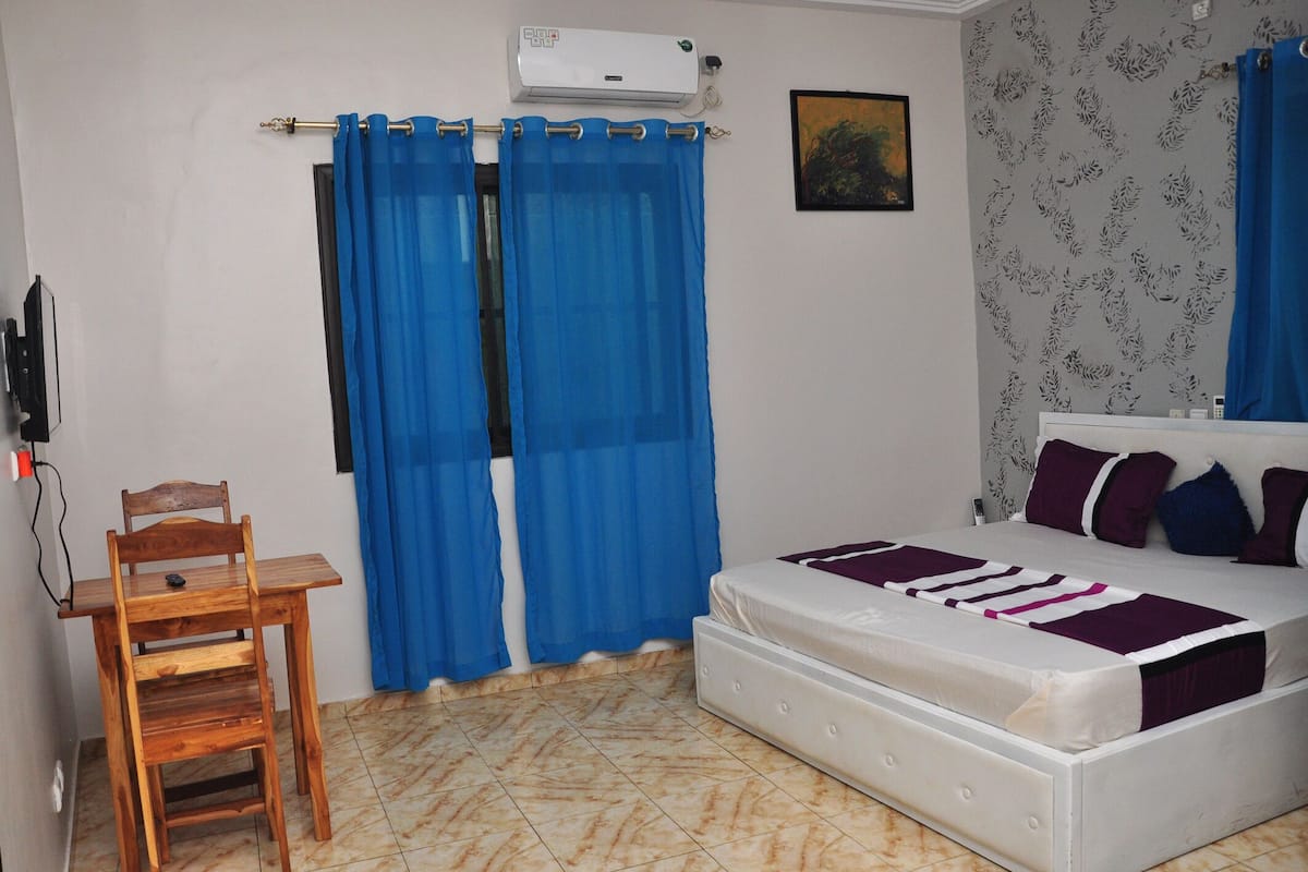 A bedroom with a bed, a chair, a window with blue curtains, an air conditioning unit, and a patterned wall.
