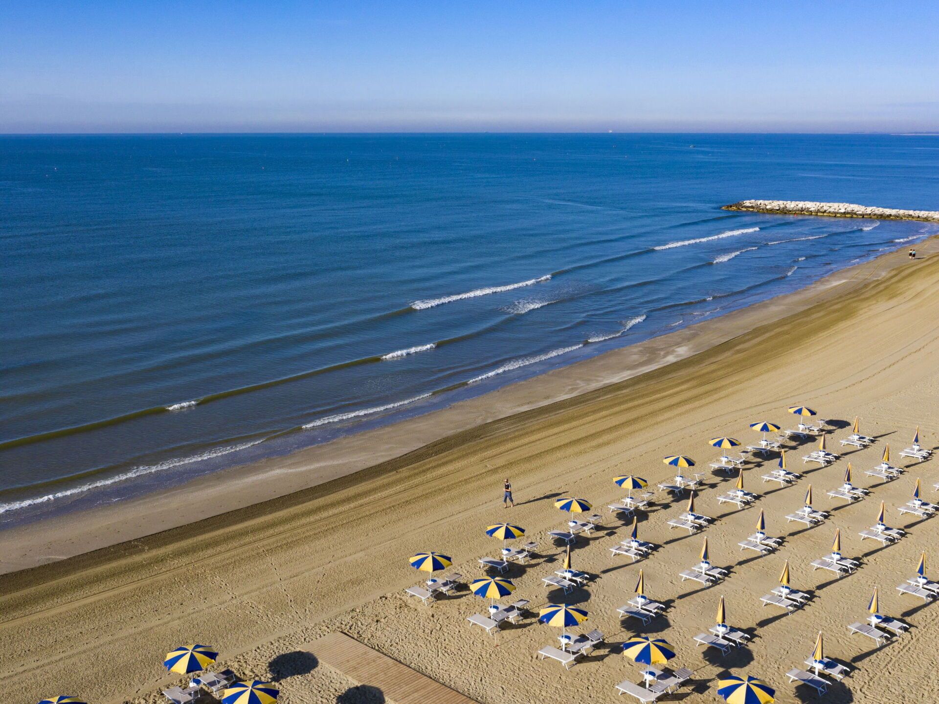 A beach with rows of sun loungers and umbrellas, a rocky breakwater, and calm ocean waves.