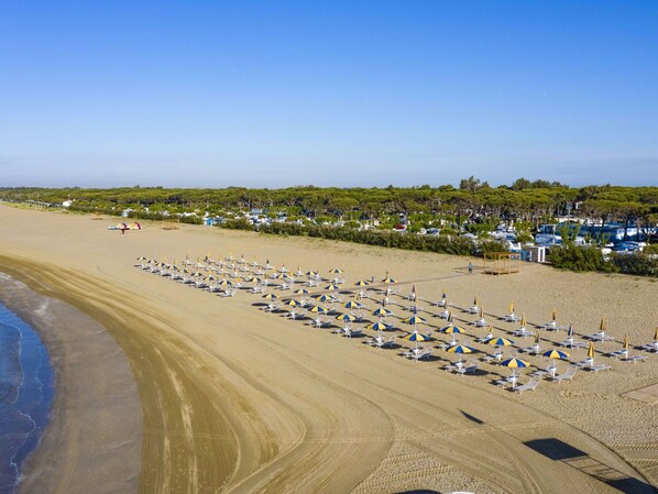 A beach with rows of sun loungers and umbrellas, a clear sky, and a parking area with cars and boats in the background.